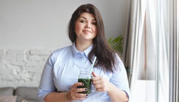Health, nutrition, dieting and detoxication concept.indoor shot of happy young brunette plus size woman with chubby cheeks holding green detox smoothie or shake, keeping vegetable diet to loose weight